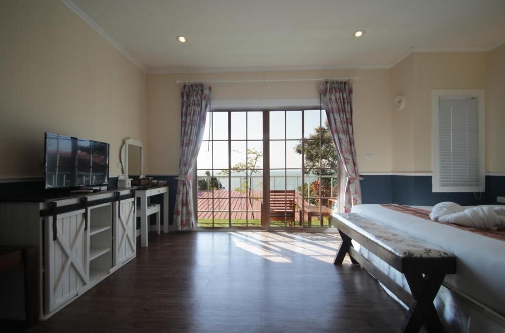 Quarto de hotel com cama, escrivaninha com televisão e uma grande janela com cortinas.