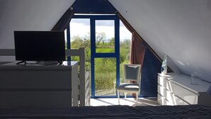 A bedroom with a TV on a dresser, a chair by the window, and a view of greenery outside. - TY LOUTI Cottage – Gateway to Huelgoat and Monts d’Arrée (Plouyé)