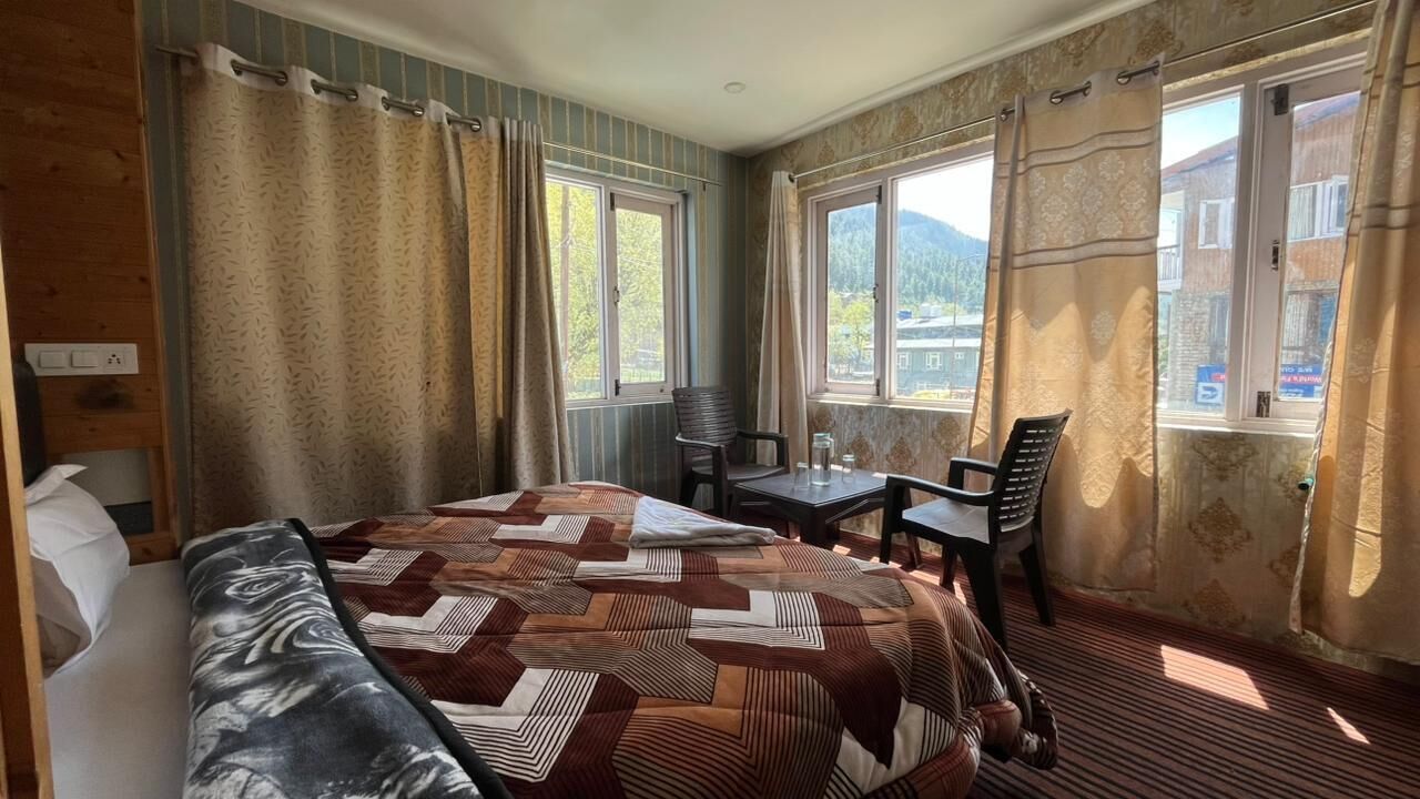 A room with a bed, a table, two chairs, and large windows with curtains.