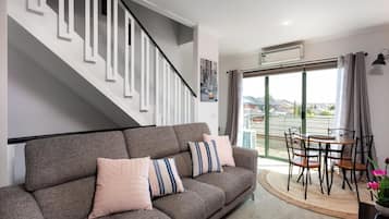 A modern living room with a grey sofa, striped pillows, a dining table with chairs, a staircase with white railings, and a large window with curtains.