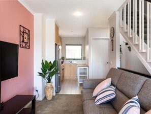 Un salon avec un canapé, un téléviseur et une plante. On y trouve également un coin cuisine avec un réfrigérateur et une fenêtre. Un escalier est visible au fond.