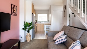 A living room with a sofa, a TV, and a plant. There is a kitchen in the background with a window. - Snug 2-bed Townhouse Near Bustling Shops (Melbourne)