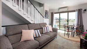 A modern living room with a grey sofa, striped cushions, a dining table with chairs, and a staircase with white railings. - Snug 2-bed Townhouse Near Bustling Shops (Melbourne)