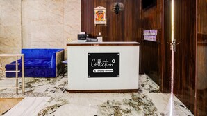 Ein Hotel-Rezeptionsbereich mit Marmorboden, einem Empfangstresen mit der Aufschrift 'Collection Company-Serviced', einem blauen Sofa, einer Holzwand mit Uhren und einer Tür mit einem Schild.