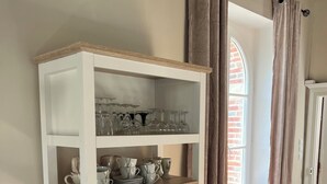 Ein weißes Regal mit Glaswaren und Geschirr, ein Fenster mit Vorhängen und ein Boden aus Ziegelsteinen.