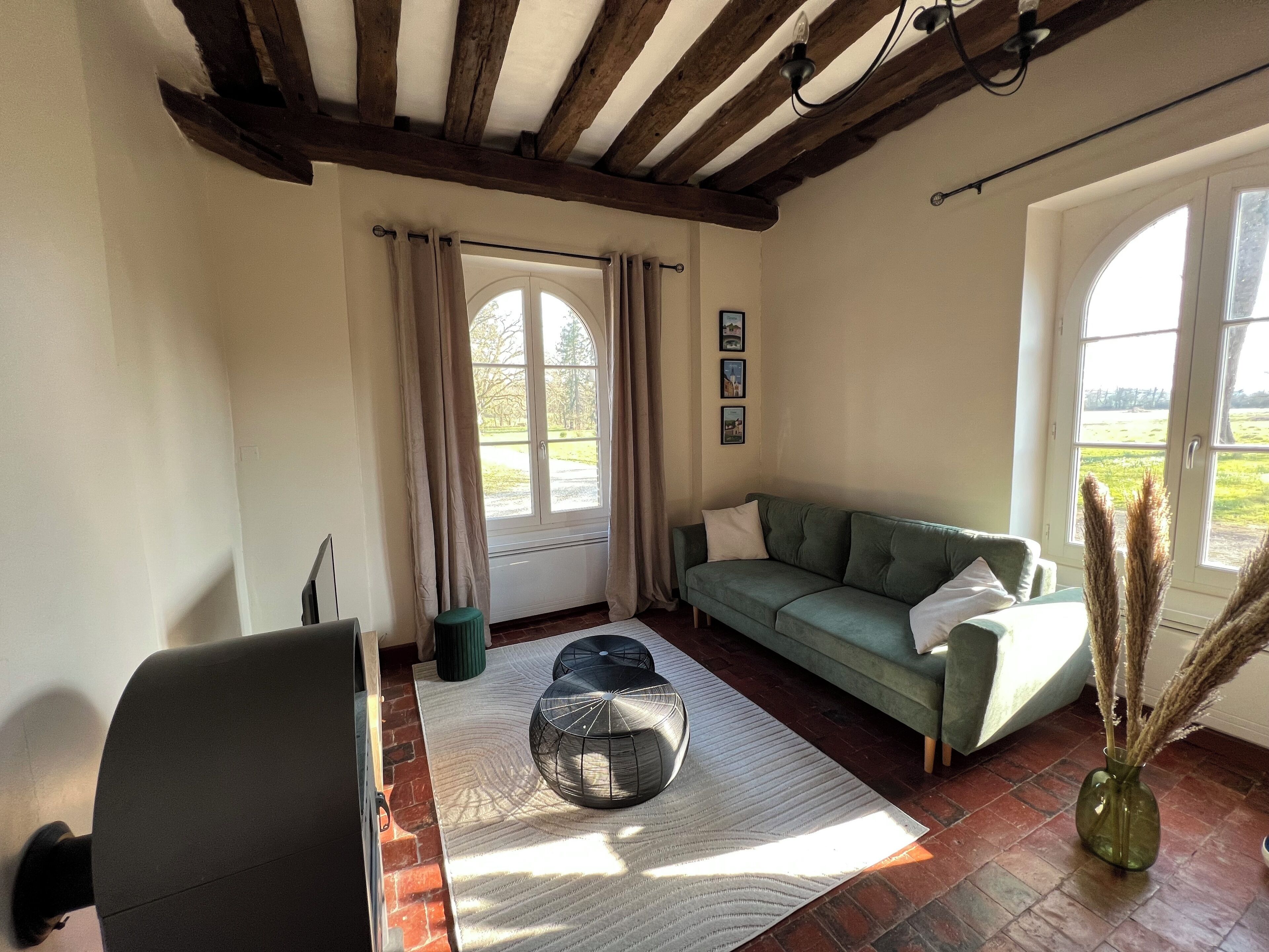 A cozy living room with a green sofa, a coffee table, and a vase with dried plants. There's a window with curtains and a view of greenery outside.