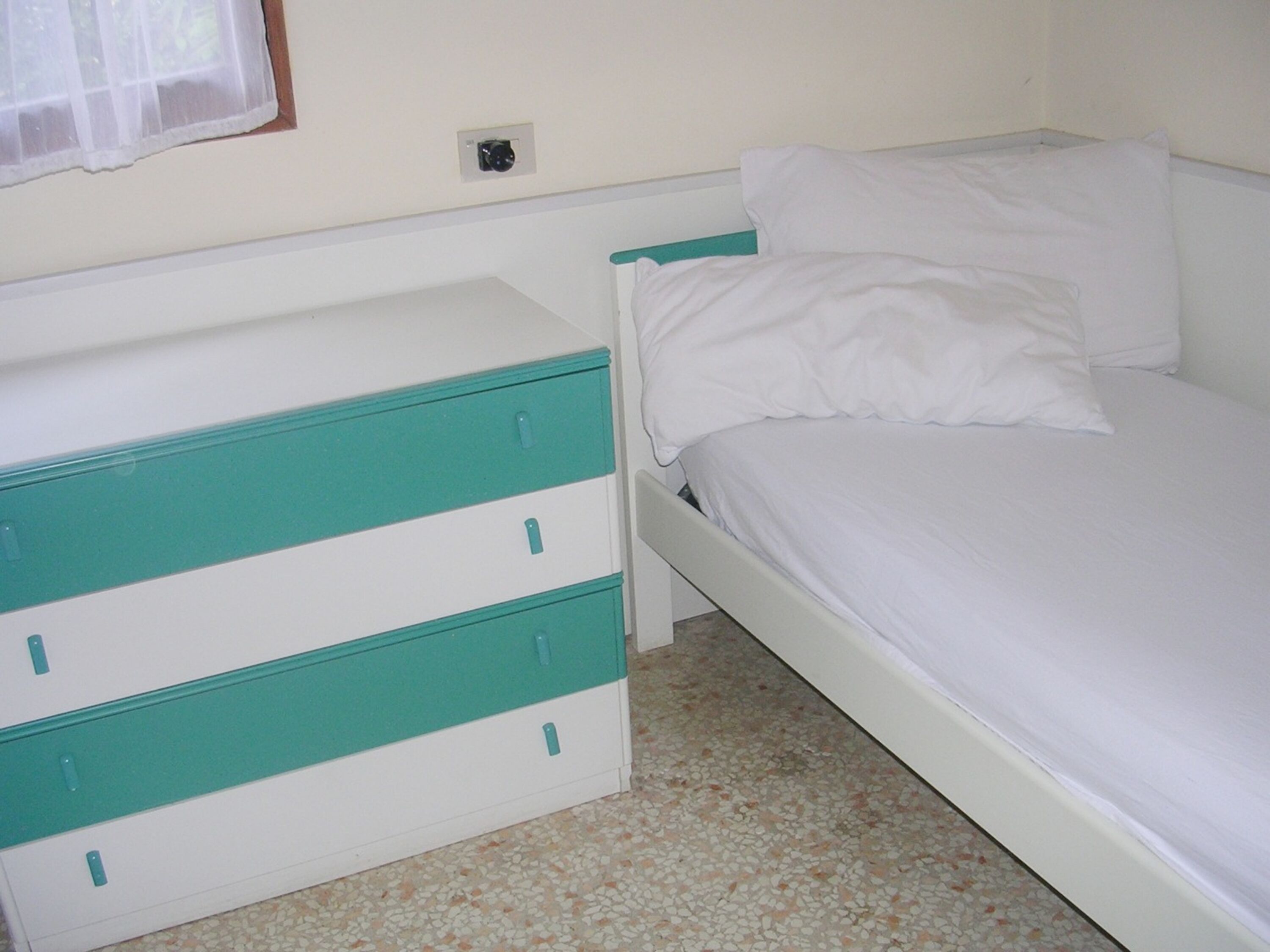 Une chambre à coucher avec un lit, une commode blanche agrémentée de touches de couleur turquoise, et un sol carrelé.