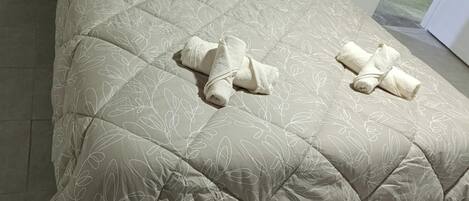 A bed with a beige quilt, two rolled towels, and a black bedside table.