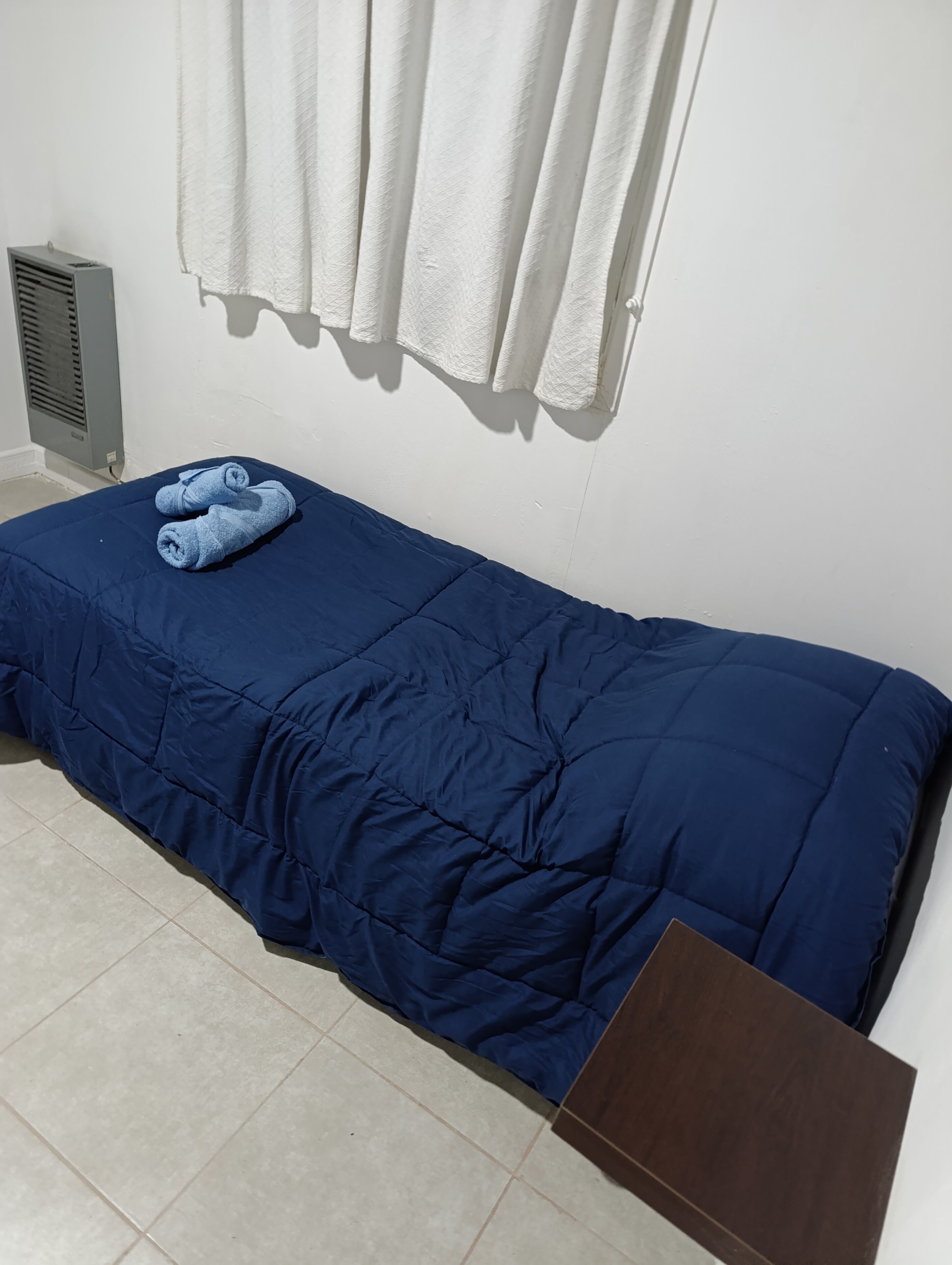 Un lit simple avec une couette bleue, une serviette blanche et une boîte marron posée sur le sol.