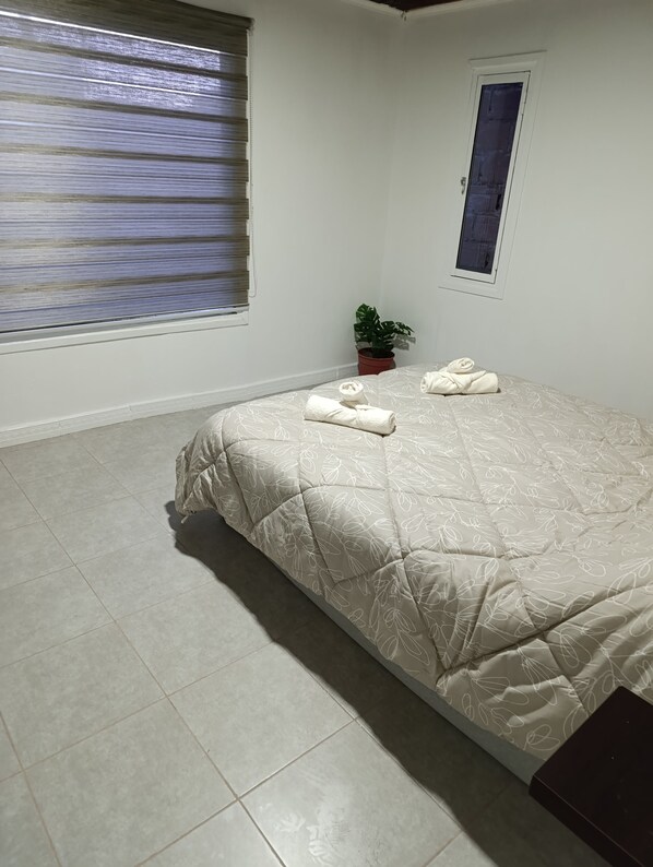 A bedroom with a bed, a potted plant, a window with blinds, and a door.
