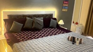 Un lit bien fait, avec une couvre-lit à carreaux, une tête de lit et plusieurs oreillers. On y trouve une table de chevet avec une lampe et un petit objet décoratif.