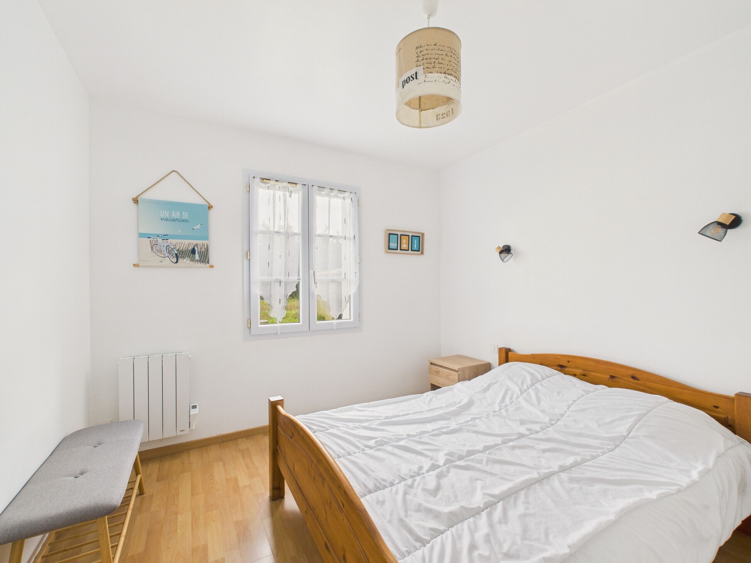 A bedroom with a wooden bed, a bench, a radiator, a window with curtains, a wall-mounted light, and a hanging light fixture.