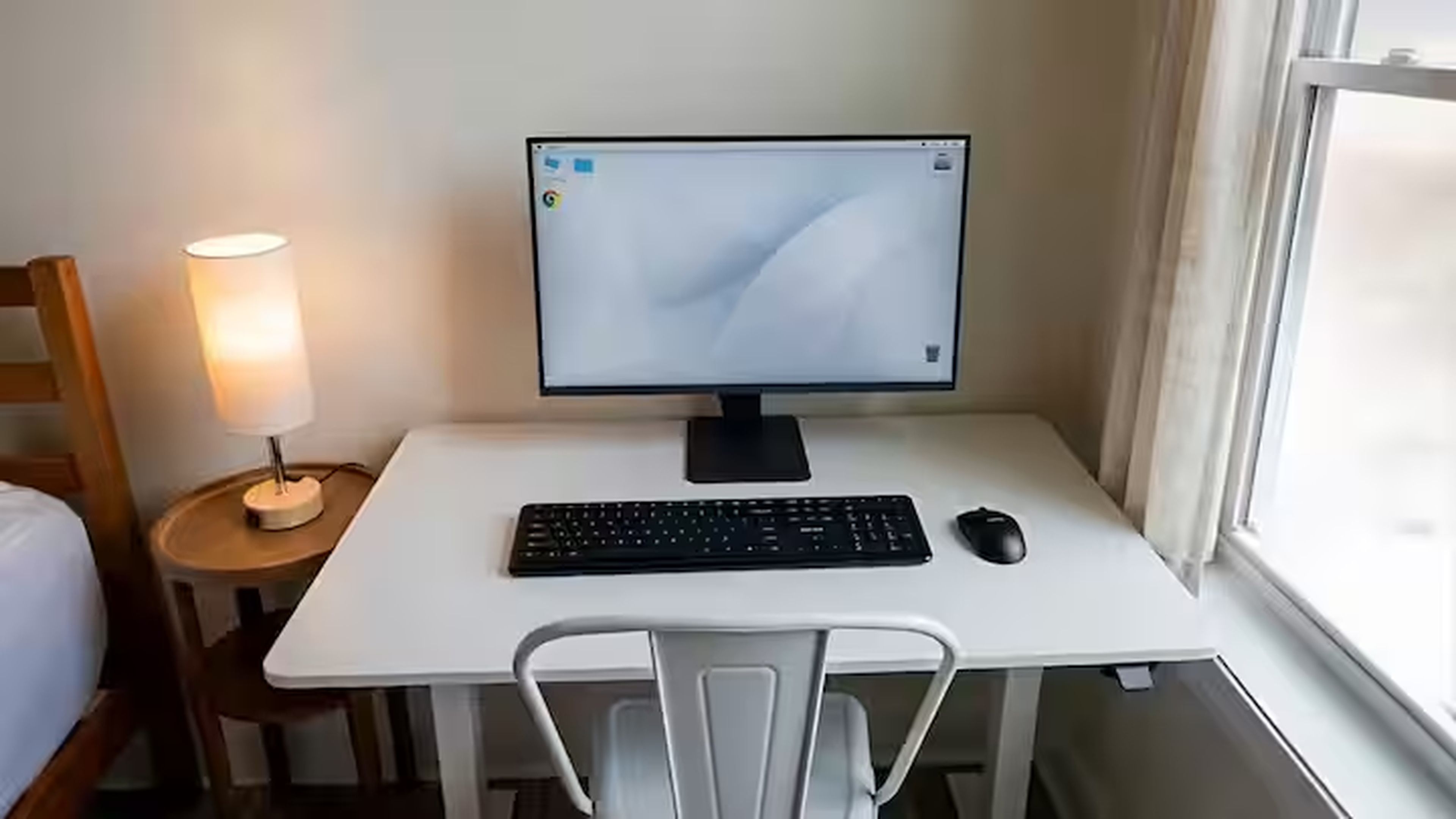 A hotel room with a desk, a computer, a keyboard, a mouse, a chair, and a bedside table with a lamp.
