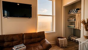 A living room with a brown leather couch, a flat-screen TV mounted on the wall, a window with a view, and a storage unit with folded towels. - Chic Wilmington Retreat—Brand-New, Central Location, Pets Welcome, Fast WiFi! (Wilmington)