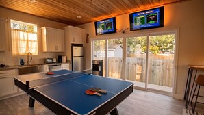 A ping pong table in a kitchen with a TV displaying a game, wooden cabinets, and a sliding glass door leading outside. - Chic Wilmington Retreat—Brand-New, Central Location, Pets Welcome, Fast WiFi! (Wilmington)