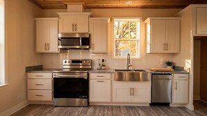 Una cocina con armarios blancos, electrodomésticos de acero inoxidable, un ventanal con vistas a árboles y un techo de madera.