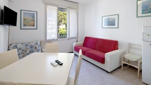 Ein Zimmer mit einem weißen Tisch, einem roten Sofa, einem an der Wand befestigten Fernseher und einem Fenster mit Blick auf Grünflächen.