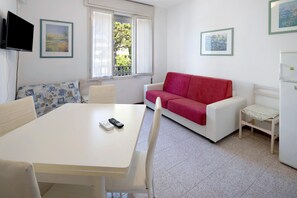 Una habitación con una mesa blanca, un sofá rojo, un televisor montado en la pared y una ventana con vistas a zonas verdes.