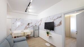 Een moderne woonkamer met een grijze bank, een eettafel met stoelen, een kleine koelkast, een aan de muur bevestigde televisie en een potplant.