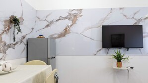 Een moderne kamer met een marmeren wand, een flatscreen televisie bevestigd boven een klein tafeltje, een zithoek met een tafel gedekt voor twee personen, en een bloempot op tafel.