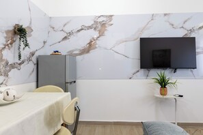 A modern room with a marble wall, a flat-screen TV mounted above a small table, a dining area with a table set for two, and a potted plant on the table. - Cozy 1-bedroom apartment in charming Saranda with AC (Saranda)