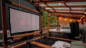 Un entorno exterior acogedor con una terraza de madera, una pantalla grande que muestra 'NETFLIX' y un jacuzzi.