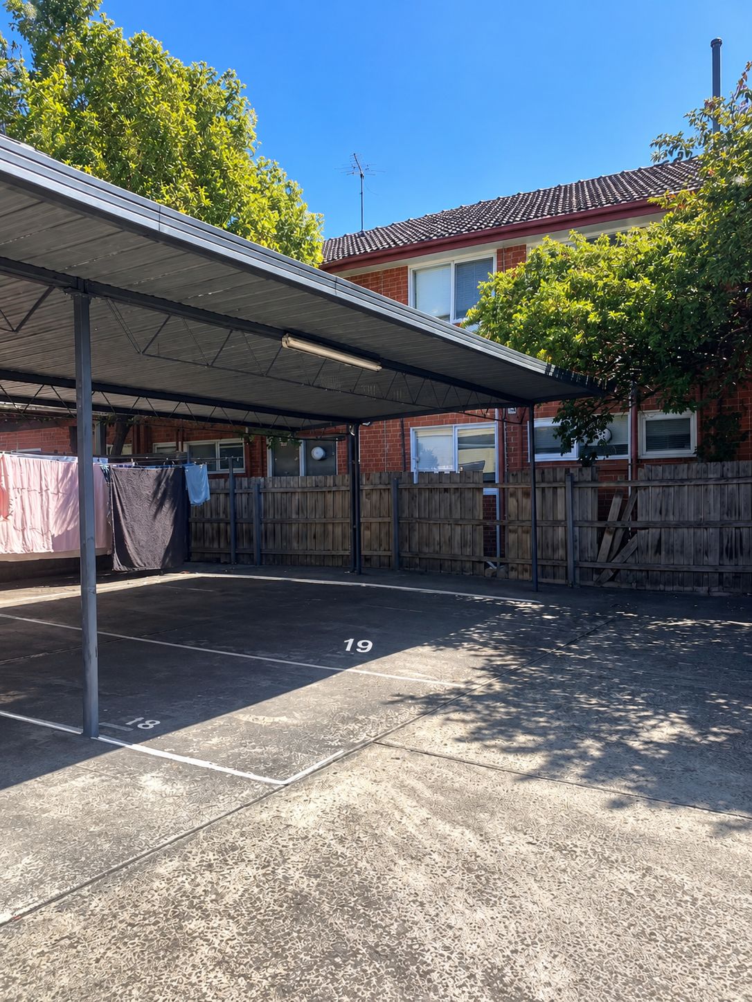 Un espace de stationnement couvert, doté d’un toit en métal, d’une clôture en bois et d’un bâtiment en briques en arrière-plan.