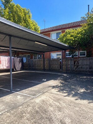 Un espace de stationnement couvert, doté d’un toit en métal, d’une clôture en bois et d’un bâtiment en briques en arrière-plan.
