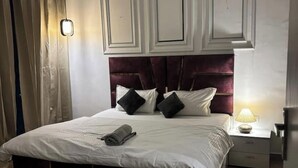 A neatly made bed with white linens and a rolled towel, a dark red upholstered headboard, and a bedside lamp on a nightstand. - Premium 14th-Floor Studio with Balcony & Stunning City Views (Noida)