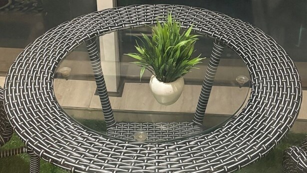 A round outdoor table with a woven metal frame and a glass top, surrounded by a green lawn. - Premium 14th-Floor Studio with Balcony & Stunning City Views (Noida)