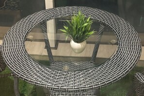 A round outdoor table with a woven metal frame and a glass top, surrounded by a green lawn. - Premium 14th-Floor Studio with Balcony & Stunning City Views (Noida)