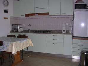 A kitchen with white cabinets, a stove, a sink, and a dining table with a floral tablecloth. - Gardened Villa in Quiet Area Near the sea (San Michele al Tagliamento)