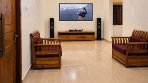 Ein Wohnzimmer mit Holzboden, einem an der Wand befestigten Flachbildfernseher, zwei hölzernen Sofas, einem Holzschrank und einem Standventilator.