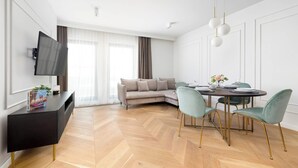 En moderne stue med sofa, spisebord og stole. Der er et fladskærms-tv på væggen, et konsolbord med et magasin, og en dekorativ plante.