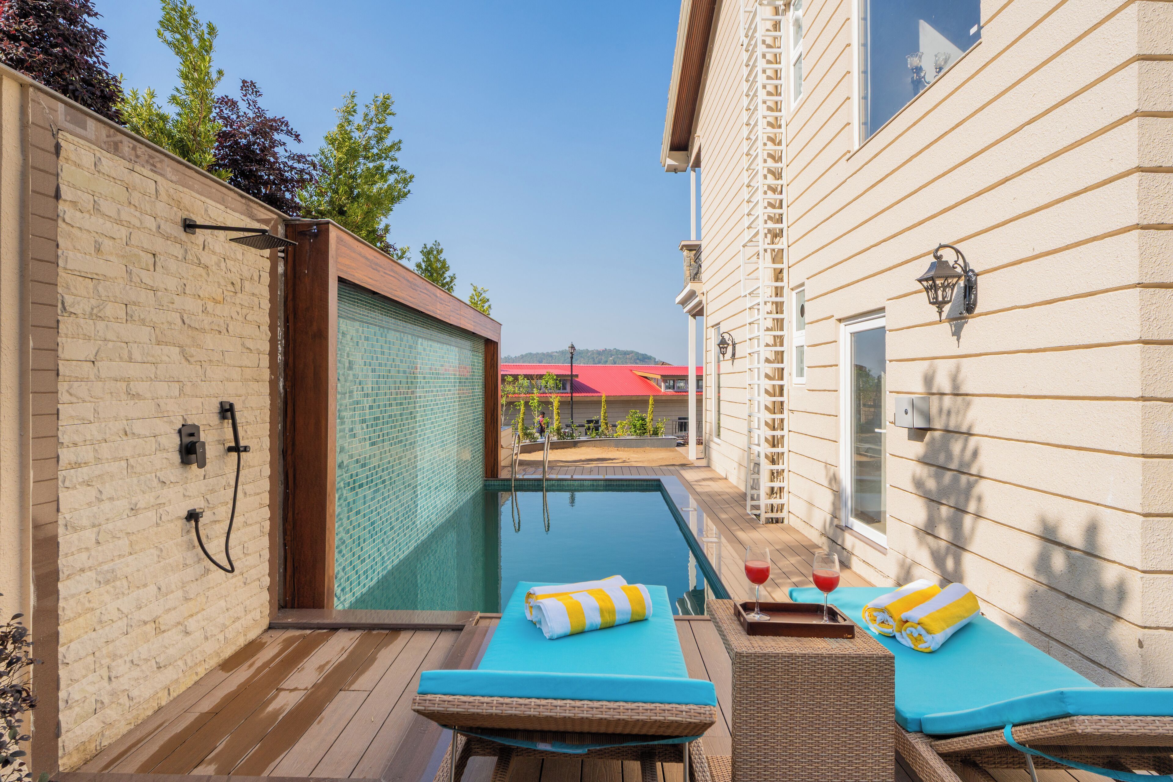 Un área junto a la piscina con sillones de descanso, una piscina cubierta con cristal y vista a un edificio de techo rojo al fondo.