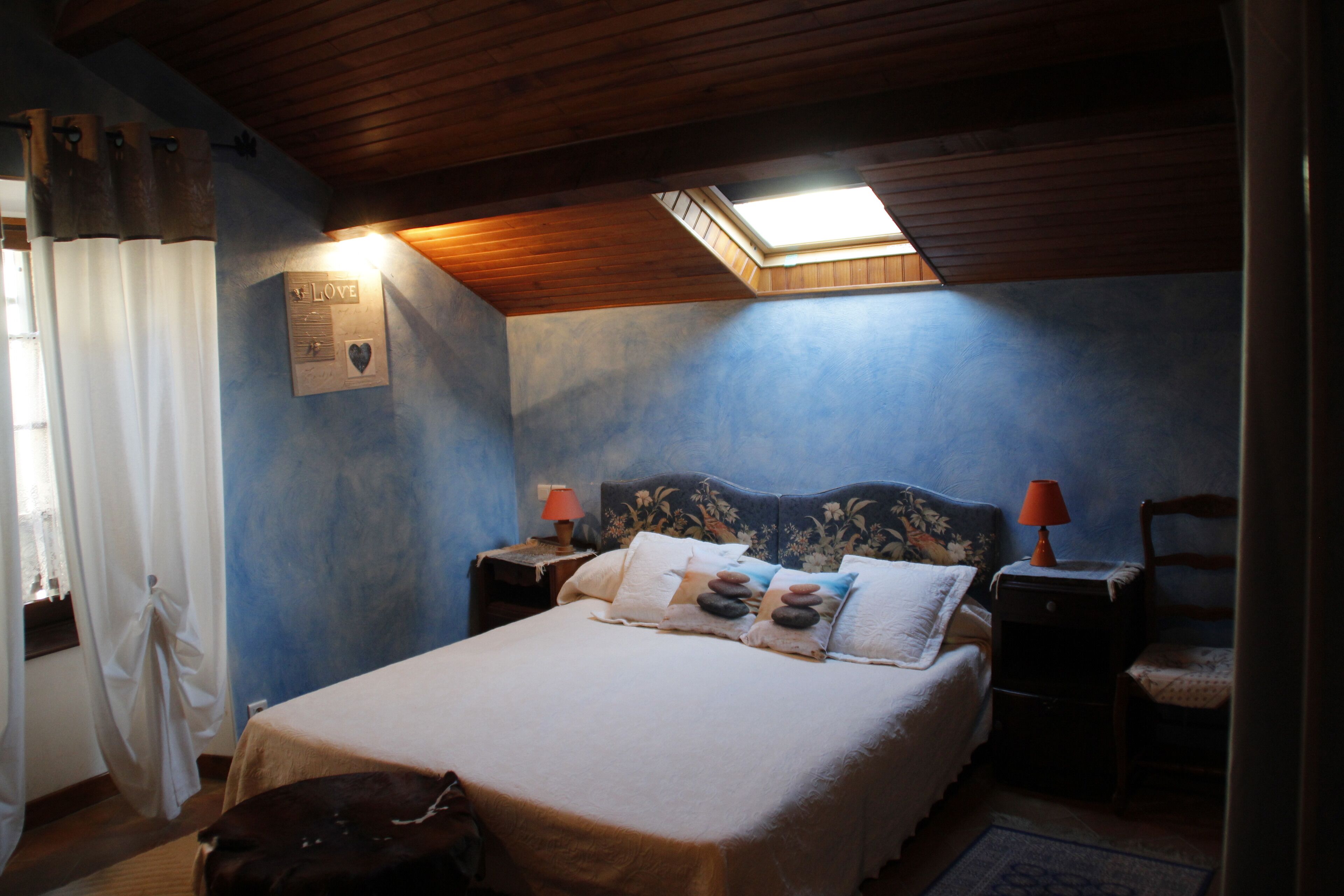 Ein Schlafzimmer mit einem Bett, zwei Nachttischen, einem Sessel, einem Fenster mit Vorhängen und einem Oberlicht.
