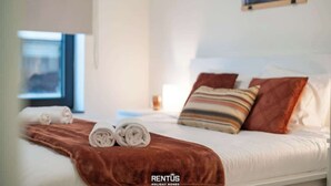 En snyggt bäddad säng med en brun filt och hoprullade handdukar, ett fönster med utsikt och en tavla på väggen.