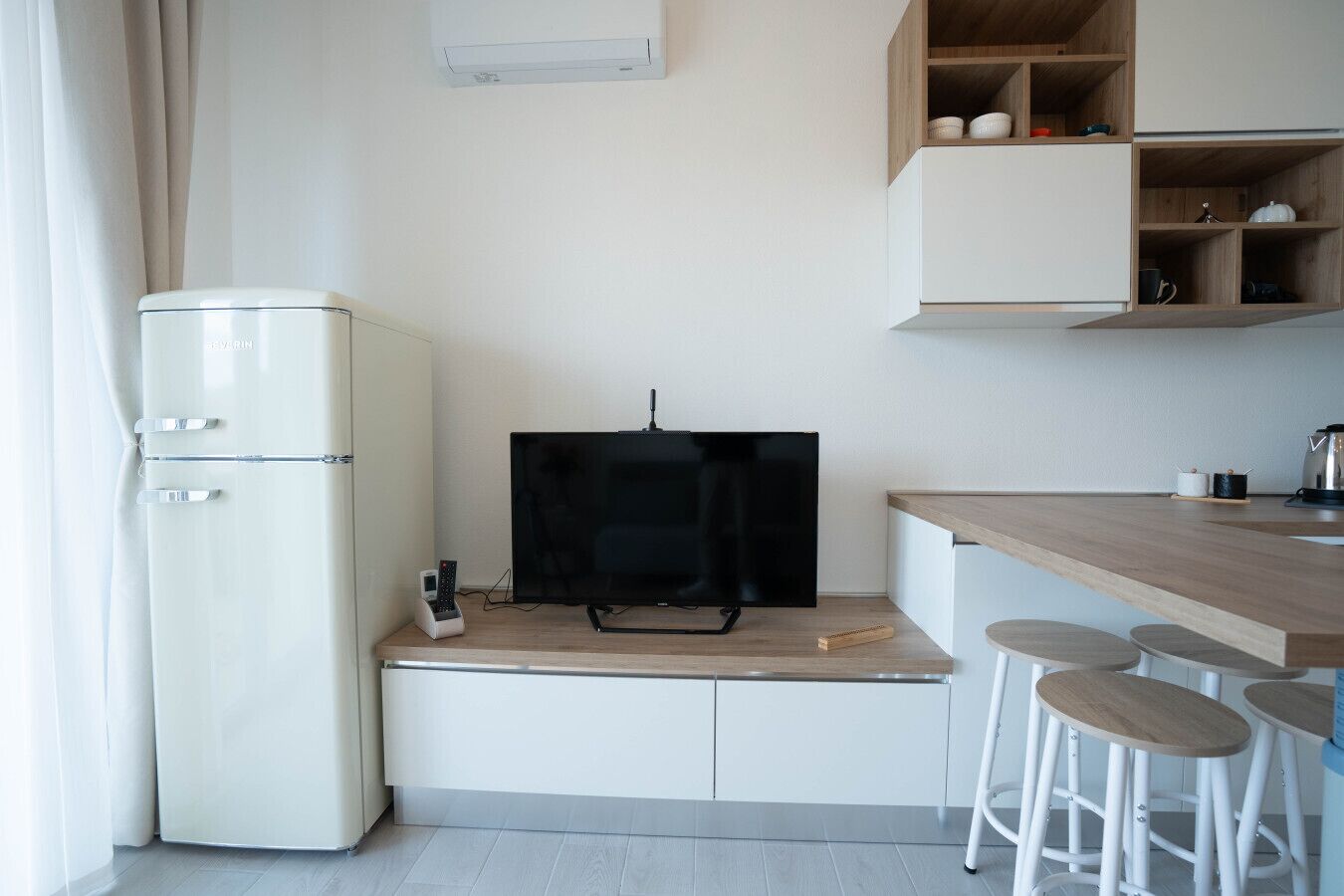 Une cuisine compacte avec un réfrigérateur blanc, un téléviseur noir posé sur un meuble en bois, et un coin bar avec des tabourets.