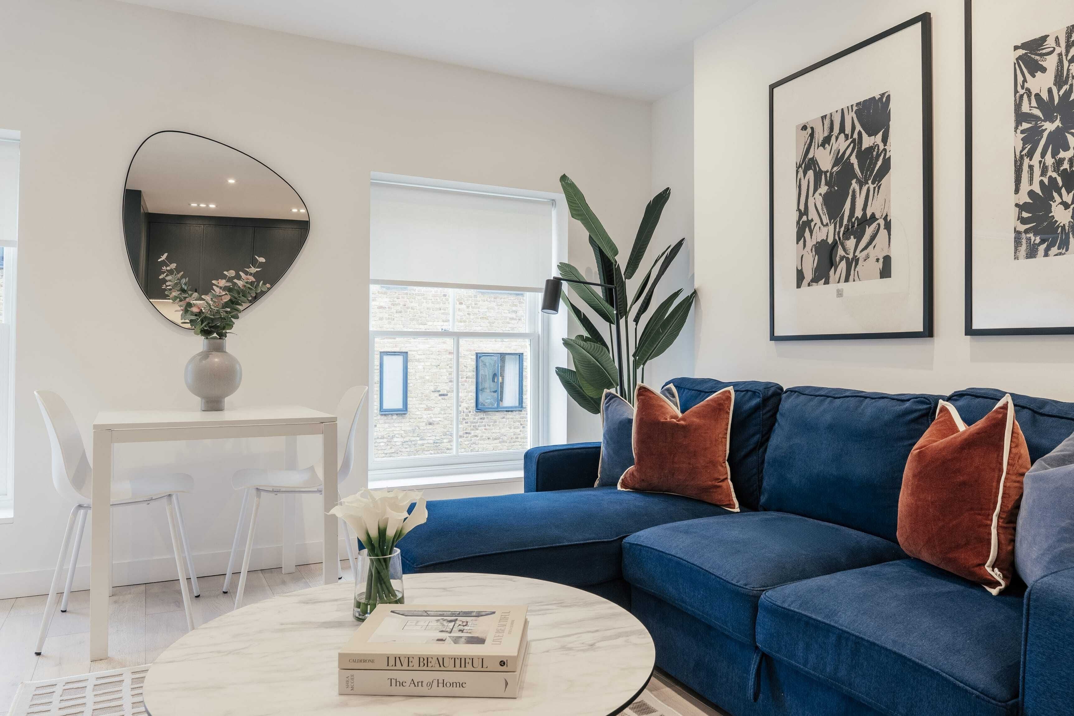 A modern living room with a blue sofa, a round coffee table, and a white dining table with chairs. There is a large mirror on the wall and framed artwork above the sofa.