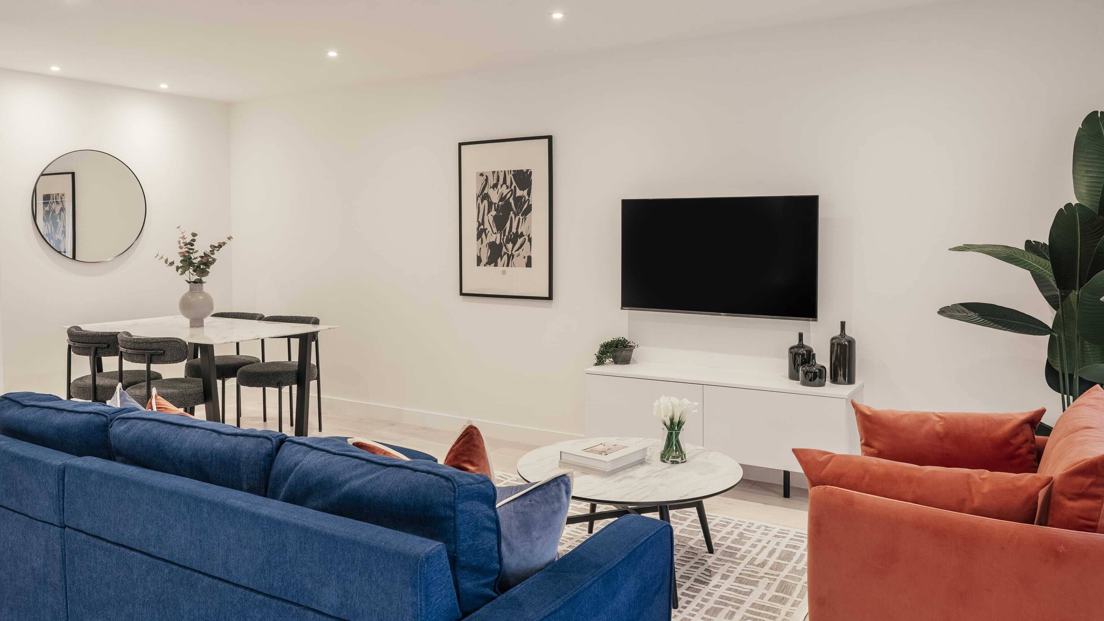 A modern living room with a blue sofa, orange armchair, and a dining area with a round table and chairs.