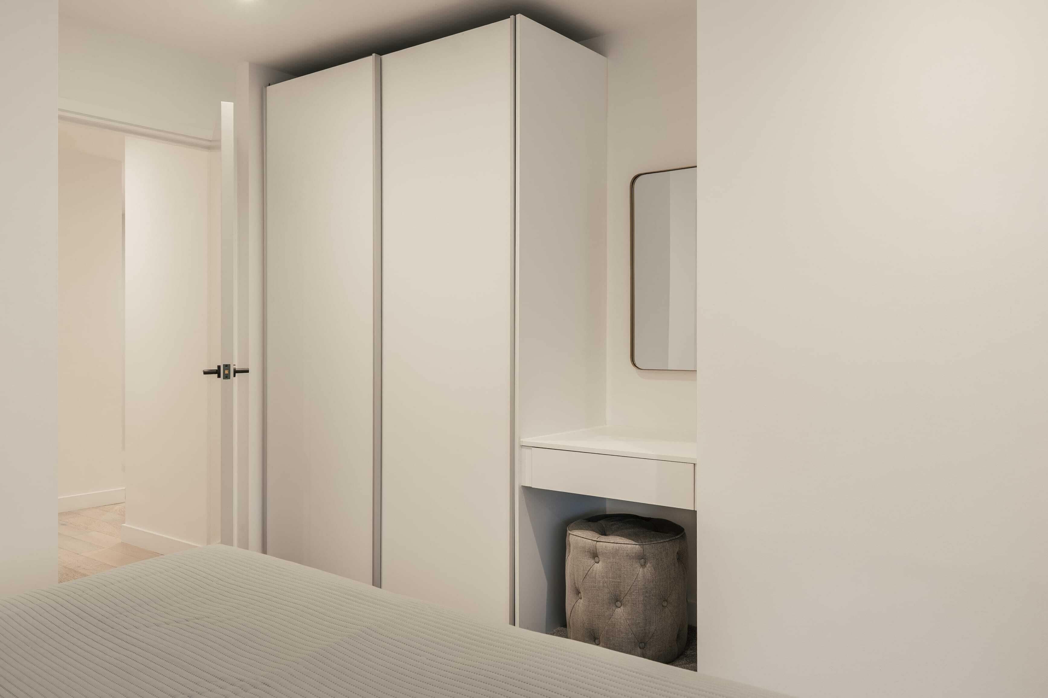 A bedroom with a bed, a wardrobe, a mirror, and a small ottoman.