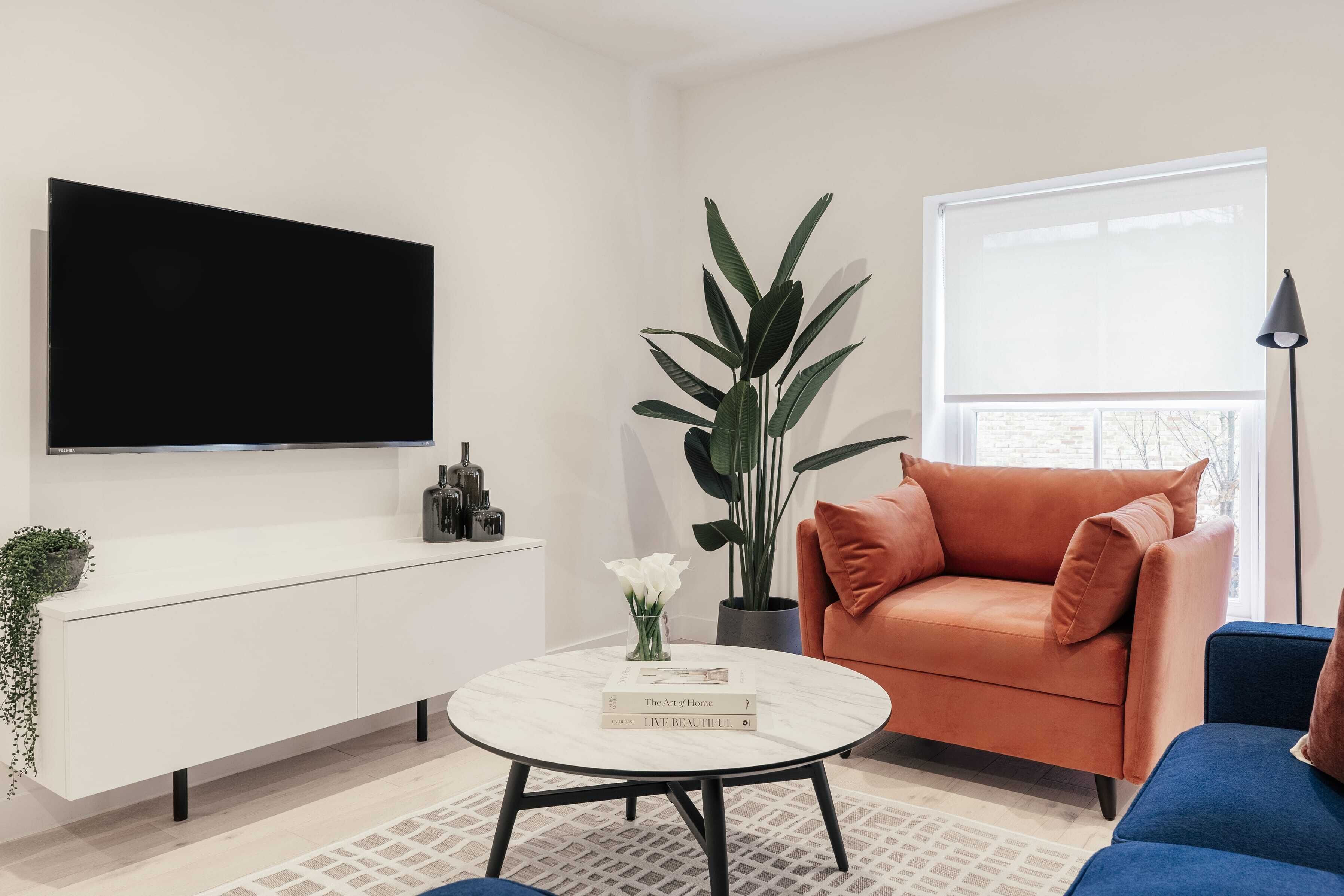 Uma sala de estar moderna com um sofá branco, uma mesa de centro redonda, uma grande planta em vaso e uma TV de tela plana na parede.