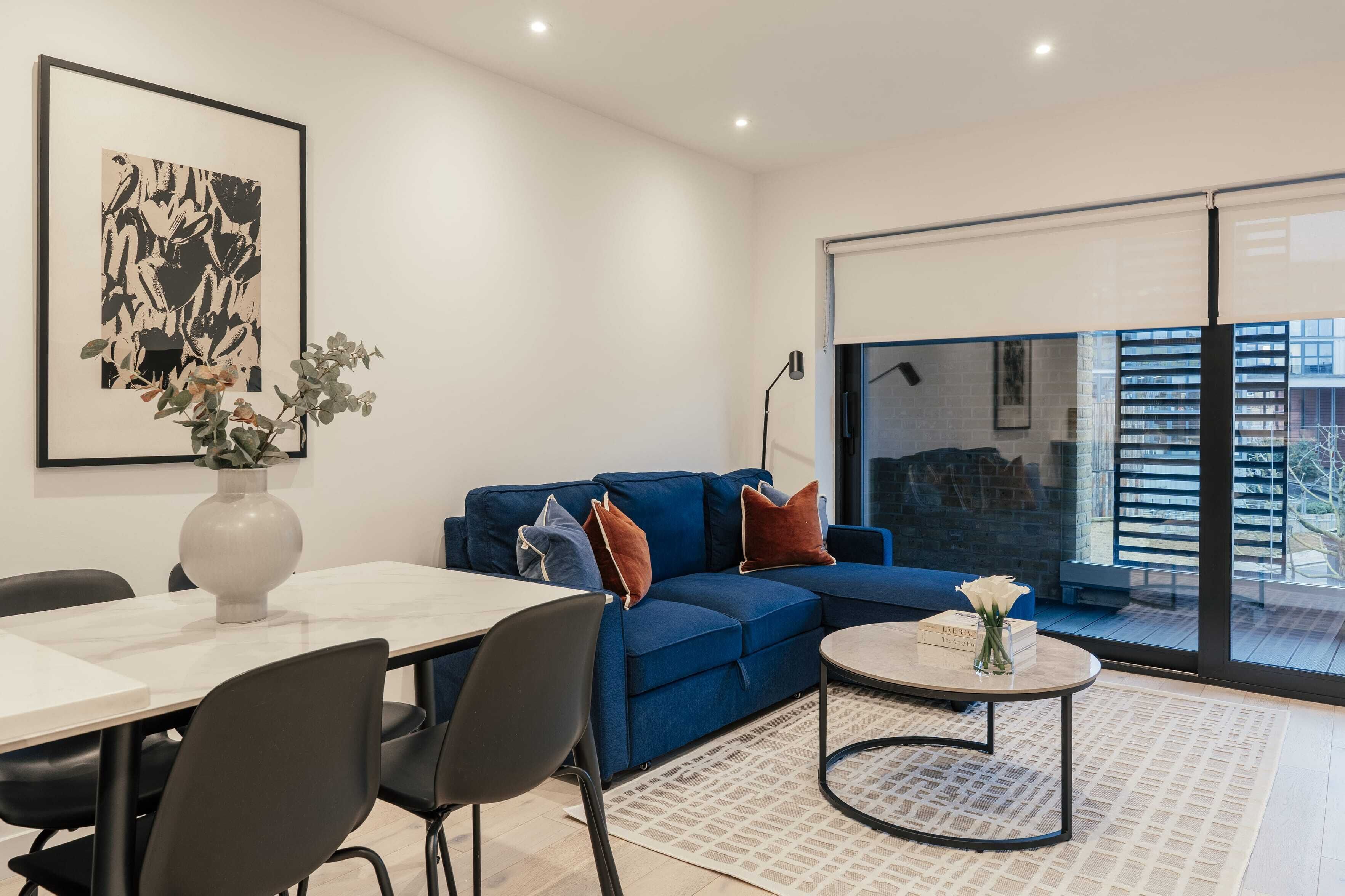 A modern living room with a blue sofa, a dining table with chairs, a round coffee table, and a large window with a view of buildings.