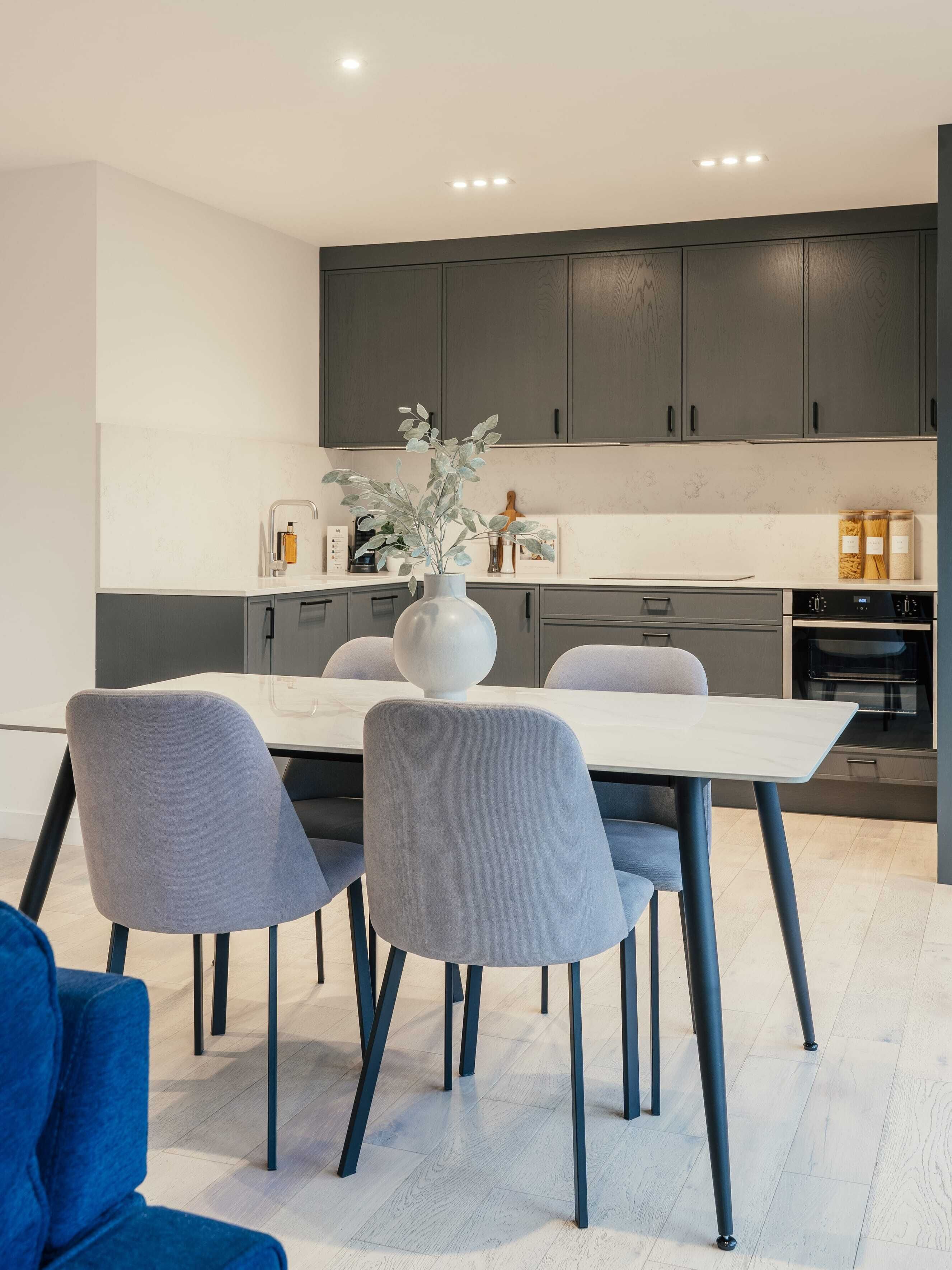 Een moderne keuken met donkere kastjes, een wit aanrecht en een eethoek met grijze stoelen en een witte tafel.