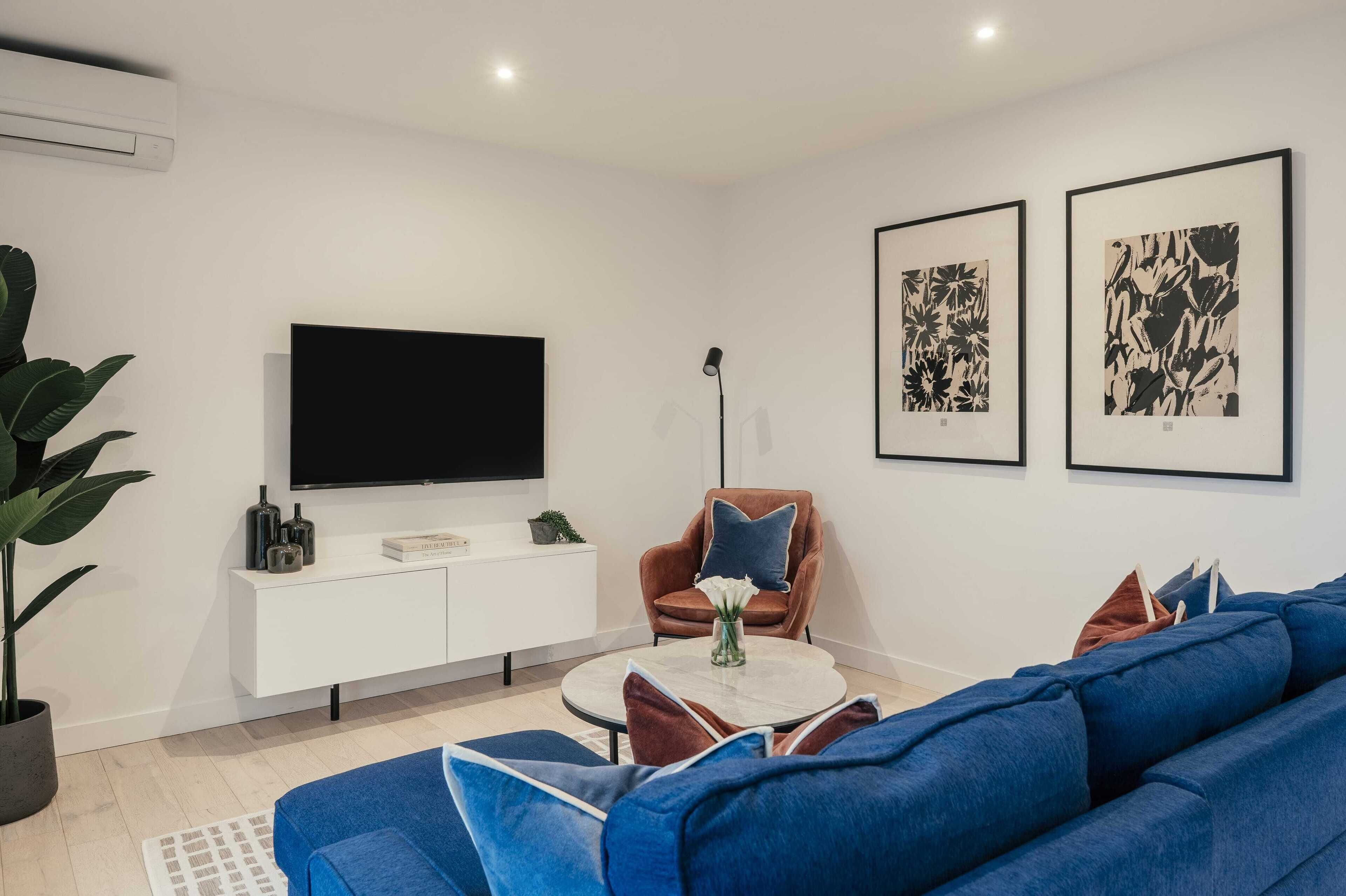 A modern living room with a large flat-screen TV, a white cabinet, a brown armchair, a blue sofa, and two framed artworks on the wall.
