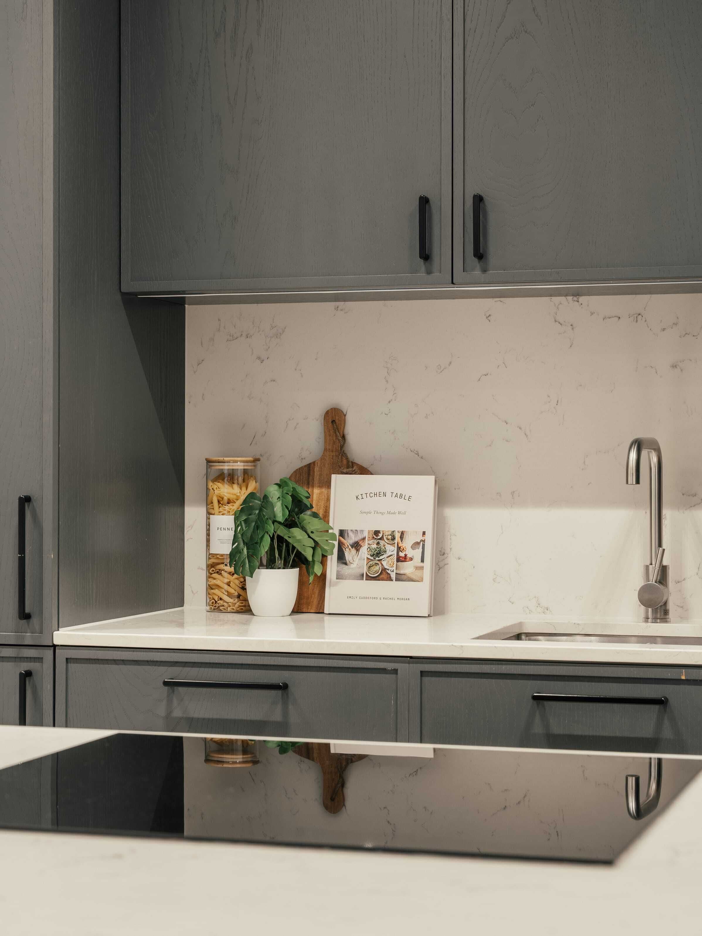 A kitchen with grey cabinets, a white countertop, a potted plant, and a cookbook titled "Kitchen Table."