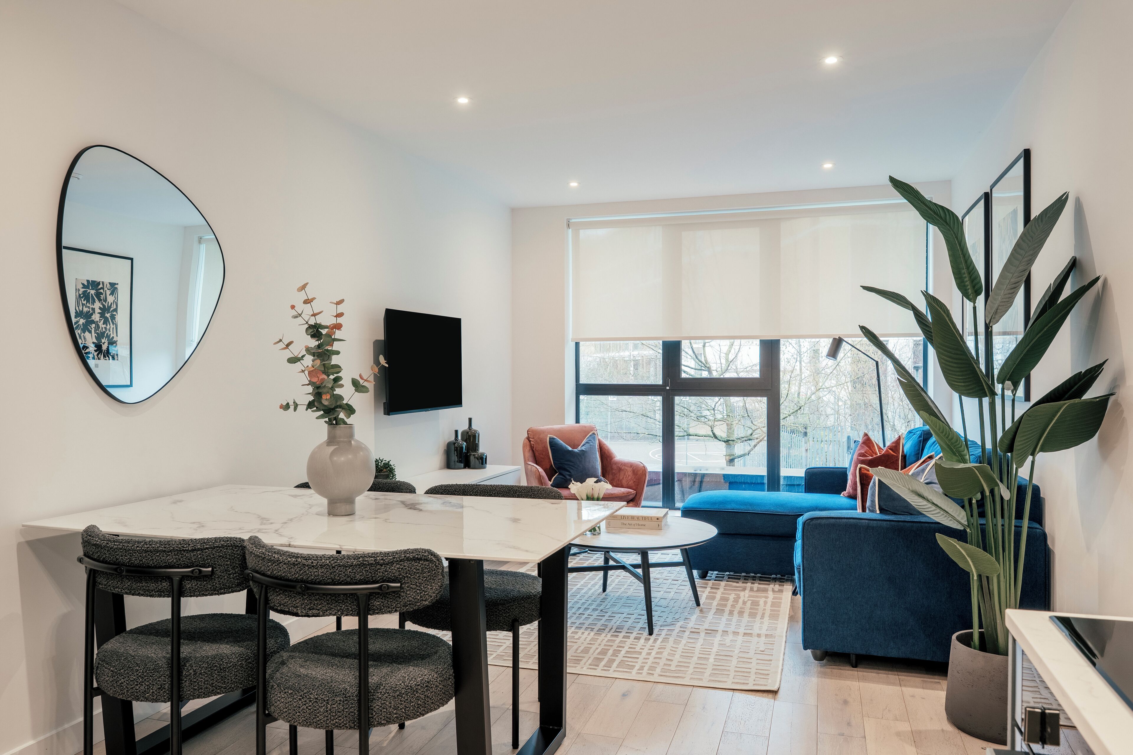 A modern living room with a round marble table, dark upholstered chairs, a blue sofa, and a large mirror on the wall.
