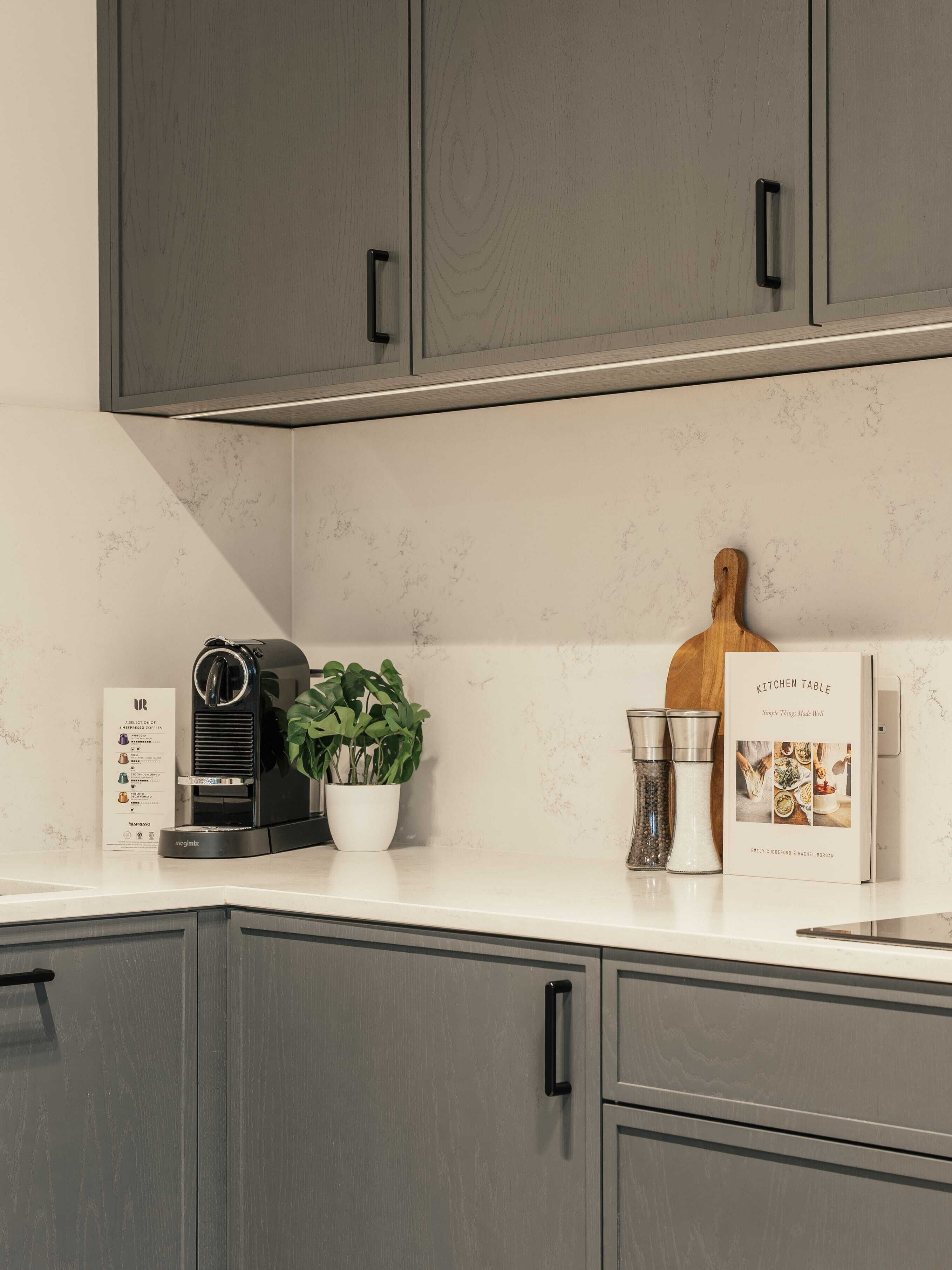 Une cuisine avec des meubles gris, une machine à café, une plante en pot, une planche à découper en bois et un livre de recettes sur le comptoir.
