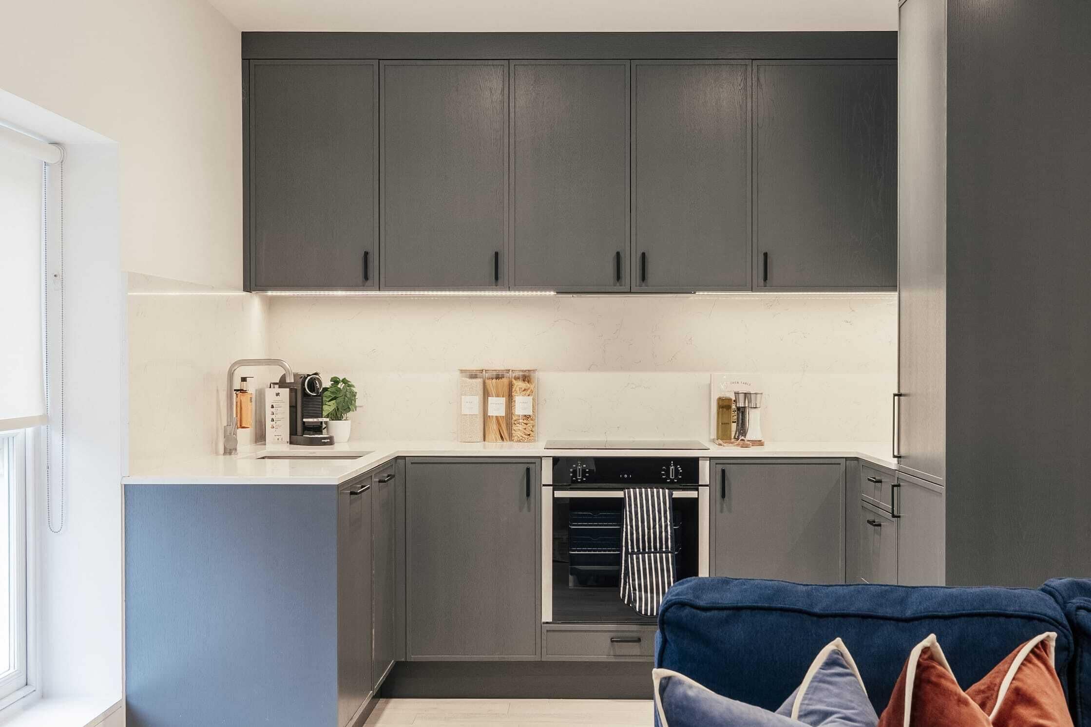 A modern kitchen with grey cabinets, a built-in oven, and a countertop with a coffee machine and a plant.