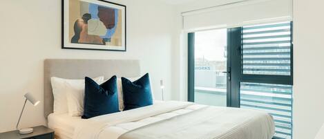 A bedroom with a bed, a nightstand, a lamp, a framed artwork, and a view through a window with blinds.